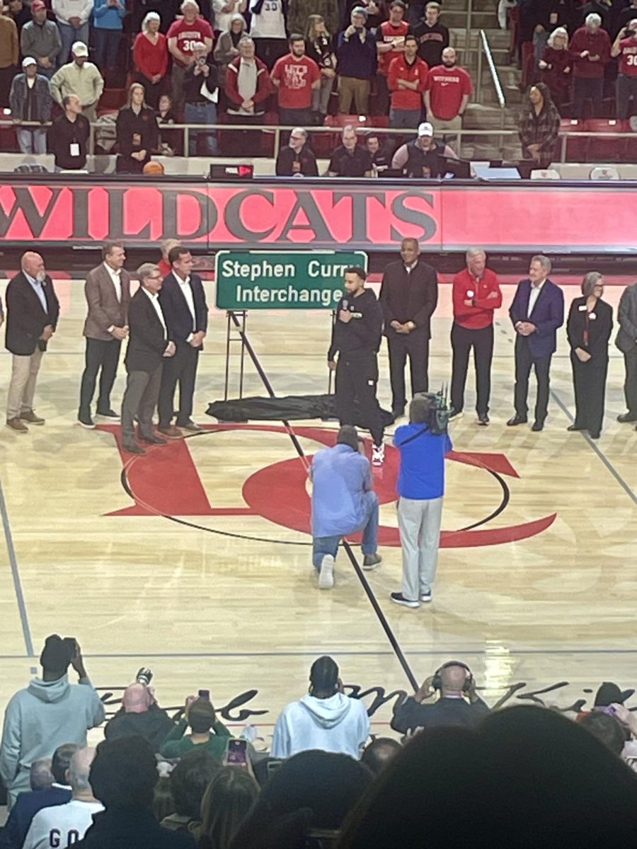 Town of Davidson and Davidson College dignitaries appear to the right and left of Davidson graduate an NBA star, Stephen Curry, at a halftime ceremony dedication of I-77’s Exit 30 being named in his honor. Holding the microphone, Steph Curry recites his speech in honor of having Interchange named after him. 

